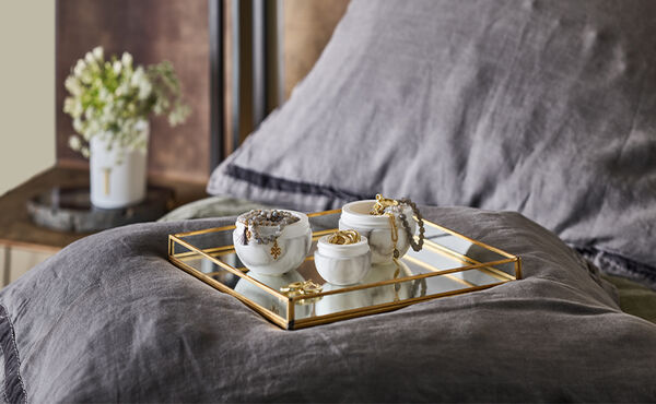 Face cream pots repurposed as jewellery holders, displayed on a gold-rimmed mirrored tray on a neatly made bed.
